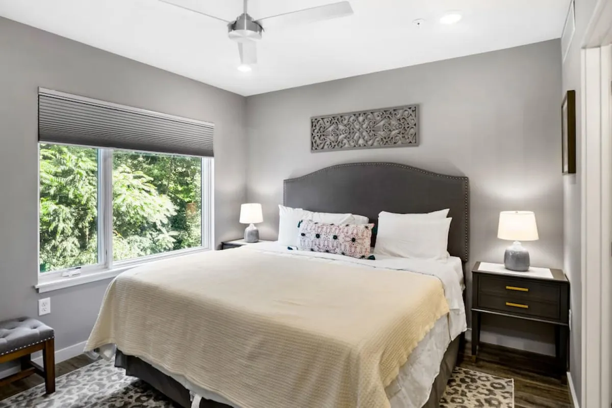 A serene bedroom featuring a bed with decorative pillows, large windows that fill the space with natural light and greenery views, and nightstands with lamps, creating a tranquil place for relaxation or reading.