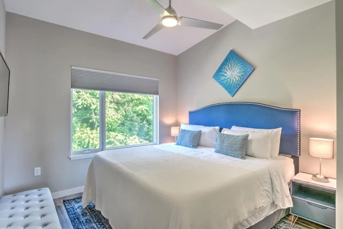 A tranquil king bedroom featuring a blue headboard, large window with a view of greenery, and tasteful wall art. The natural light streaming in creates a peaceful, inviting atmosphere, ideal for rest and reflection.