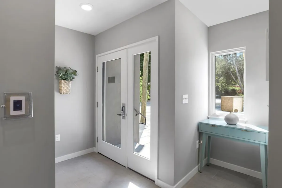 The view from inside the home looking toward the front door, with a window letting in natural light. A side table and lamp complete the entryway, evoking a sense of welcome and connection between indoor comfort and the vibrant city just outside.