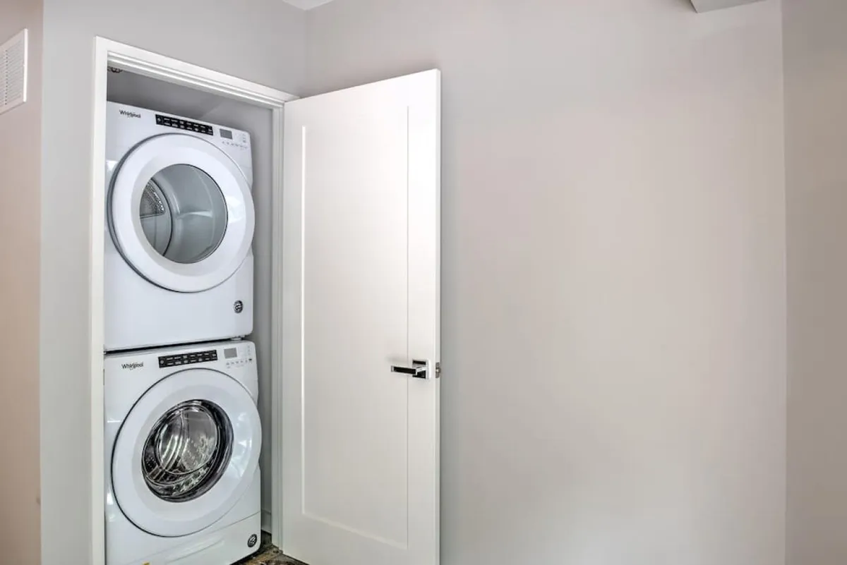A stacked washer and dryer are neatly positioned against clean white walls with a modern door nearby. The setup is compact, bright, and practical, emphasizing the convenience of in-home laundry facilities located in the garage area.