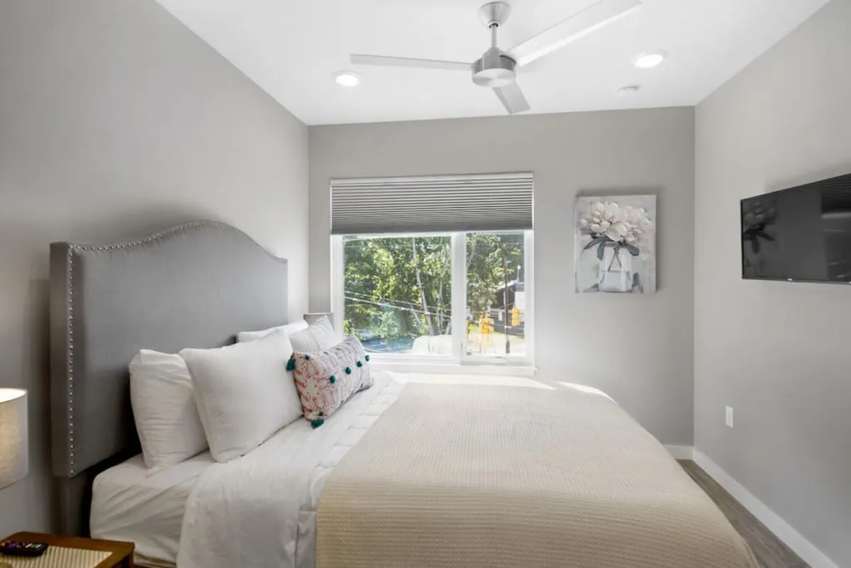 A relaxing bedroom with a bed adorned with decorative pillows, a large window letting in plenty of natural light, tasteful wall art, and a TV mounted on the wall for cozy evenings.