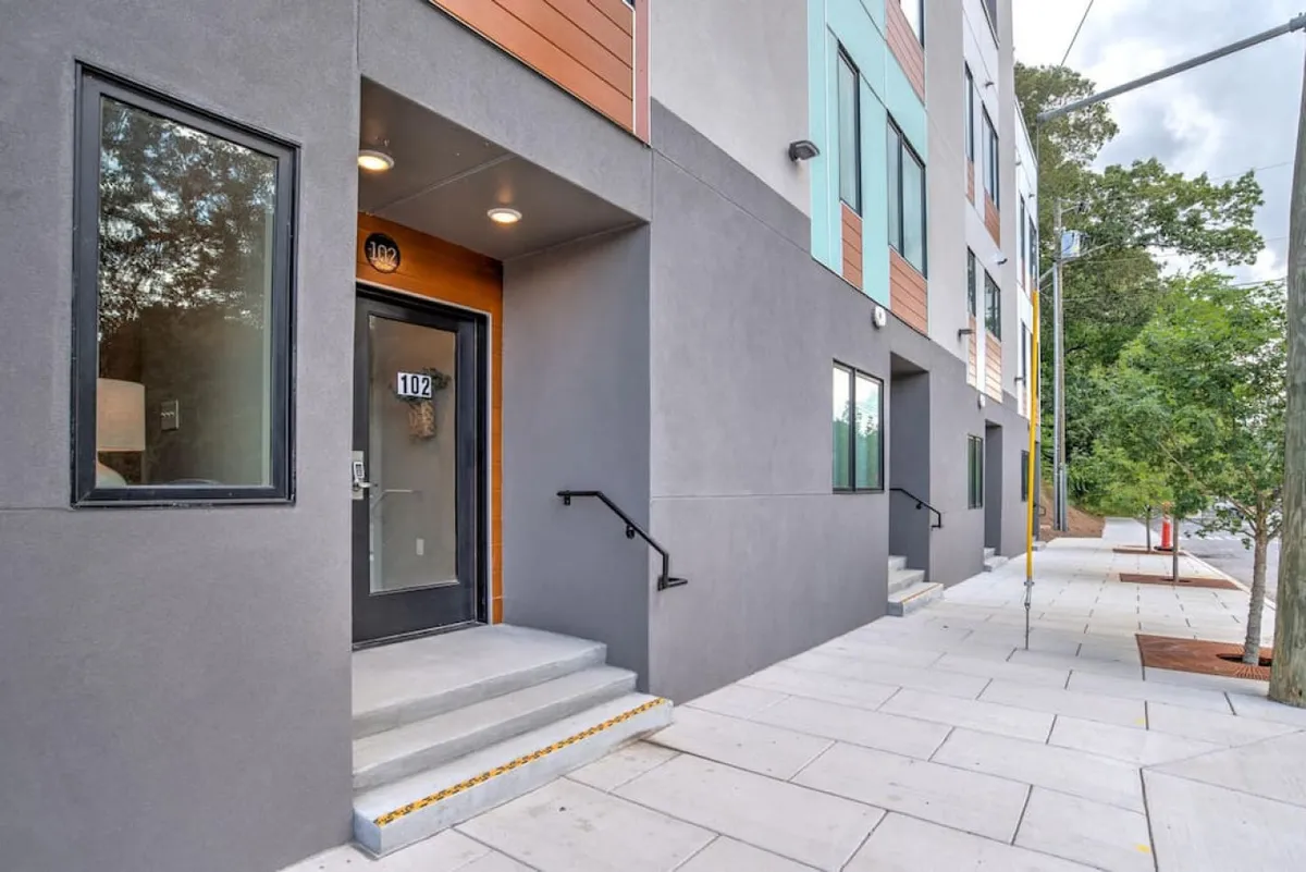 A modern exterior with a welcoming entrance door, steps leading up, and surrounding greenery. The view suggests a sense of arrival and highlights security features that might include exterior cameras, in addition to the inviting first impression.