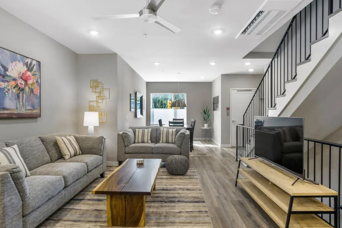 A contemporary living room with a plush sofa adorned with cushions, a sleek wooden coffee table, a TV stand with a large television, and stylish artwork on the wall. The dining area can be seen in the background, visually connecting the living and dining spaces. Bright natural light fills the room, highlighting its spaciousness and inviting atmosphere.