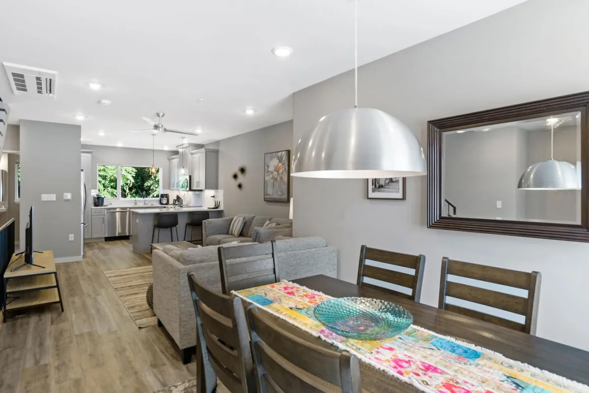 An inviting open-concept dining space with a colorful runner on the table, a pendant light fixture above, and seamless flow into an open kitchen and a cozy sofa seating area. The design encourages conversation and togetherness, with kitchen and living spaces all visible.