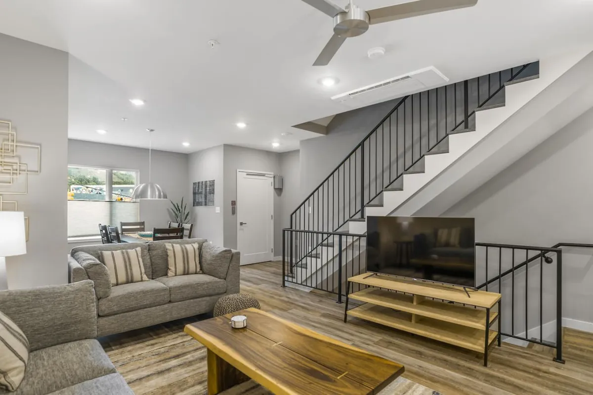 This inviting interior image highlights a spacious open-plan living room with a comfortable sofa, coffee table, TV, and a staircase leading to upper floors. The adjoining dining area is visible, all bathed in natural light, creating a cohesive space perfect for socializing, movie nights, or group gatherings.