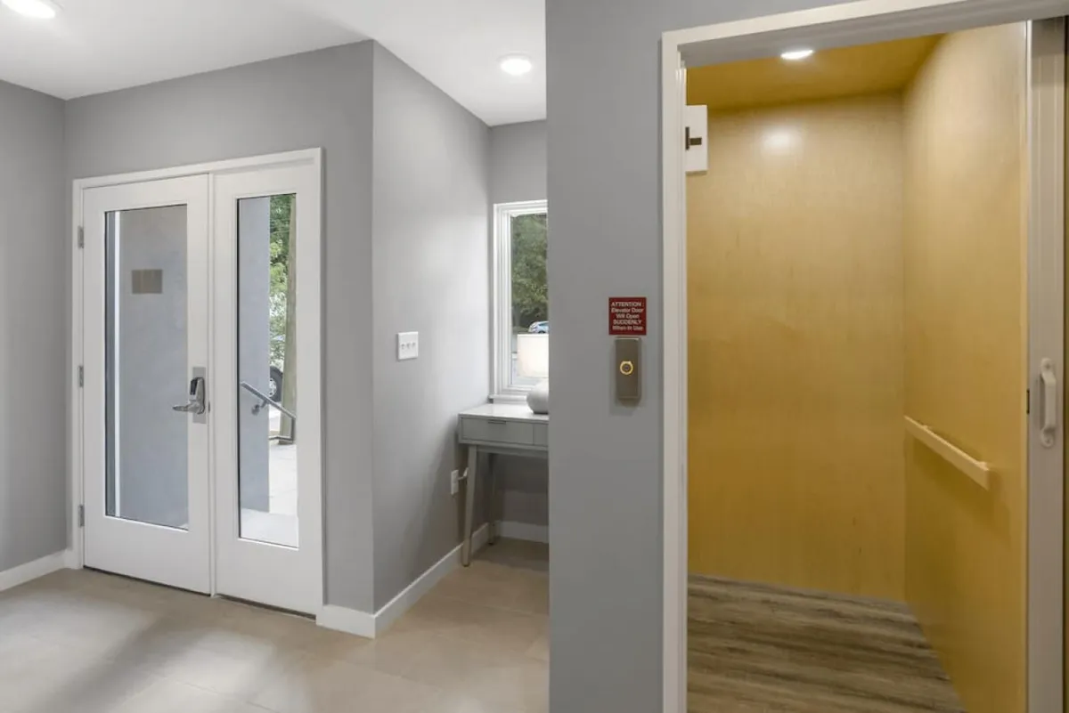 A welcoming lobby area with double glass doors, an elevator, a small table and chair, and windows that let in natural light. The space feels inviting and functional, suggesting quick and easy transitions from home to the city streets.