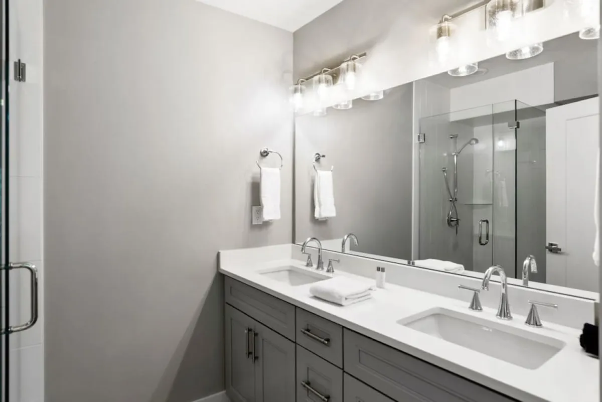 A spacious and modern bathroom with a double sink, large mirror, shower area, and elegant light fixtures. The room is bright and organized, offering a serene atmosphere for self-care routines.